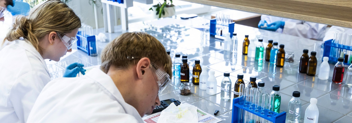 Moksleiviai chemijos laboratorijoje laiko mėgintuvėlį ir atlieka laboratorinį darbą.