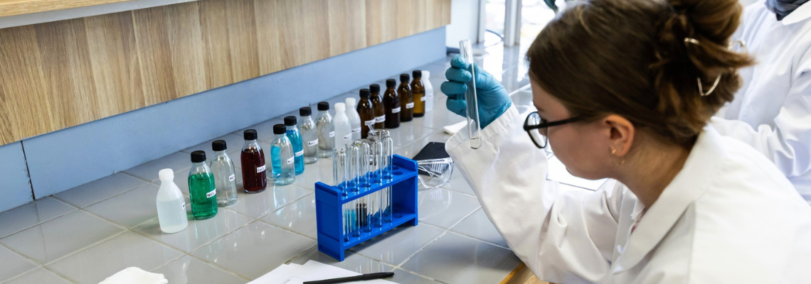 Moksleivė chemijos laboratorijoje laiko mėgintuvėlį ir atlieka laboratorinį darbą.
