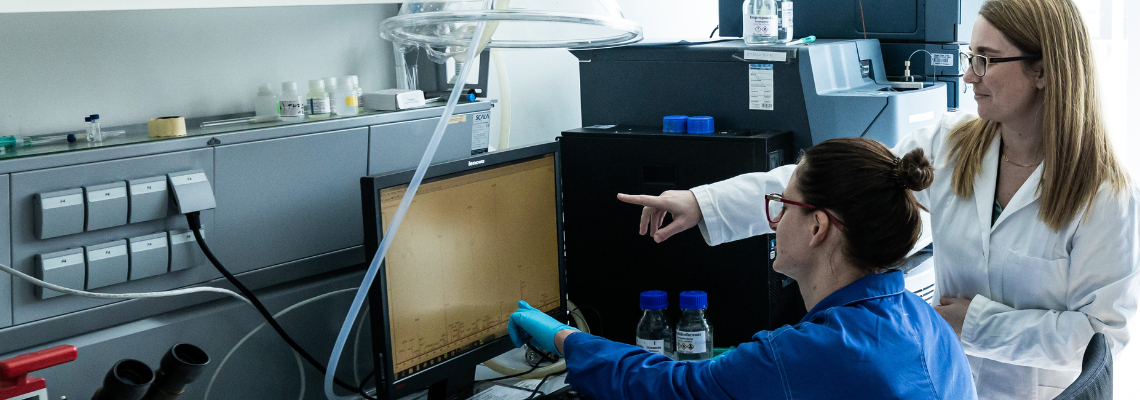 Dvi darbuotojos laboratorijoje analizuoja tyrimų rezultatus kompiuterio ekrane &scaron;alia mokslinės įrangos.
